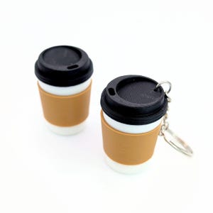 May include: Two miniature coffee cup replicas, one featuring a metal keychain attachment. Each cup is white with a black lid and a tan sleeve around the center. The items are constructed with a smooth finish and a visible textured pattern on the lids.