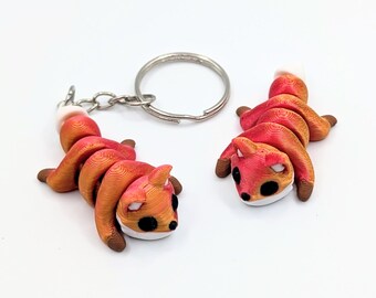 May include: Two small, articulated fox figurines with a gradient orange and red finish. One fox is attached to a silver metal keyring with a chain, while the other is separate. Both figures feature black eyes, white accents, and a layered texture.