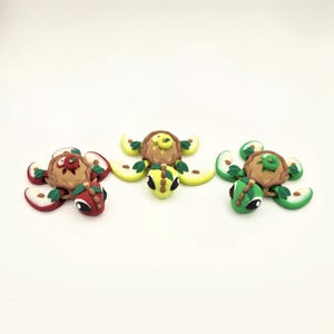May include: Three colorful polymer clay turtle figurines. Each turtle has a brown shell, apple slice flippers, and a turtle head. One turtle has red and white apple slices, another has yellow, and the last has green.