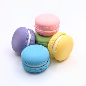 May include: A collection of five round, disc-shaped objects resembling macarons in pastel colors including purple, pink, green, blue, and yellow. Each piece features a smooth top and bottom shell with a textured, ruffled edge and a white center layer.