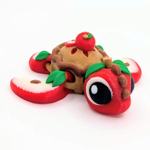 May include: A whimsical 3D-printed apple pie with a red apple on top, surrounded by red apple slices with green leaves. The pie crust is tan and brown. The apple slices have white flesh and brown seeds. The overall theme is food and dessert.