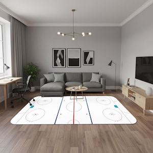 May include: A white hockey rink rug featuring blue and red markings, circles, and face-off dots. The rug is placed in a living room with a grey sofa, coffee table, and framed artwork.