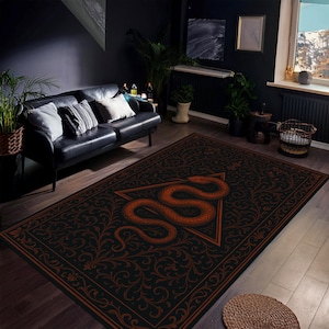 Könnte beinhalten: Ein rechteckiger schwarzer Teppich mit einem orangefarbenen Schlangenmuster in einem Dreieck. Der Teppich hat einen verzierten, floralen Rand in Orange. Der Teppich befindet sich in einem Raum mit einem schwarzen Ledersofa und Pflanzen.