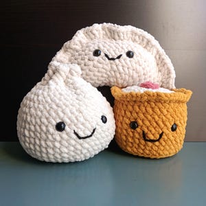 May include: Three crocheted plush toys shaped like different Asian food items. The first is a white dumpling with black eyes and a smiling mouth. The second is a yellow bun with black eyes and a smiling mouth. The third is a white dumpling with black eyes and a smiling mouth.