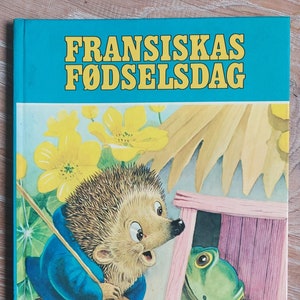 以下が含まれることがあります： 黄色い文字で「Fransiskas Fødselsdag」と書かれた青い本。表紙には、青いシャツを着て棒を持っている漫画のハリネズミが、ピンクと白の家から顔を出している緑のカエルを見ている様子が描かれている。この本は、「En Daxi Bog」というシリーズの一部で、番号は22です。