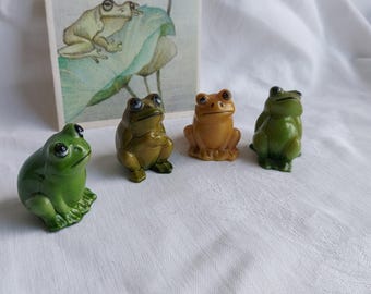 May include: Four small frog figurines, two green, one yellow, and one green with a brown back. The frogs are sitting on a white surface.