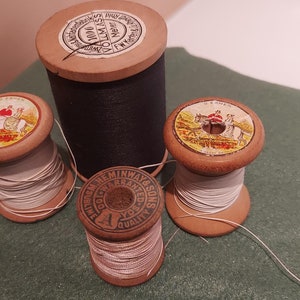 Op de afbeelding: Drie houten spoelen met draad, één zwart en twee wit. De spoelen hebben etiketten met afbeeldingen van paarden en ruiters. De zwarte spoel heeft een etiket met de tekst "1000 YARDS 200/6"