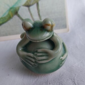 Op de afbeelding: Een klein keramiek kikker beeldje met een groene glazuur. De kikker zit met gekruiste benen en zijn handen voor zich gevouwen.