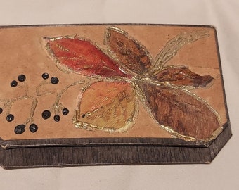 May include: A small, rectangular box with a brown leather exterior. The box is decorated with a painted design of a stylised flower with red, orange, and brown leaves. The flower is accented with gold paint. The box has a dark brown wooden base.