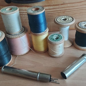 Può includere: Una collezione di otto bobine di filo di legno, alcune con etichette, tra cui "J. & J. Brock & Co.", "D.M." e "S.C.K.E.R.B.E.R.". Le bobine sono disposte su una superficie di legno, insieme a un infila-ago di metallo.
