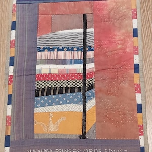 Op de afbeelding: Een kleurrijk patchwork-deken met verschillende stoffen en texturen. Het deken is omzoomd met een rand van blauwe, oranje en witte stof. De tekst "MAXIMA, PRINCES OP DE ERWT?" is geborduurd aan de onderkant van het deken.