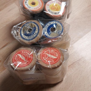 Könnte beinhalten: Eine Sammlung von acht Garnrollen, verpackt in durchsichtigem Plastik. Die Rollen sind aus Holz und haben verschiedene Etiketten mit Text in verschiedenen Sprachen. Die Etiketten sind rot, blau und weiß. Die Rollen sind in zwei Stapeln angeordnet.