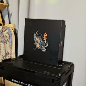 Può includere: Scatola nera con un drago e il carattere cinese "Kai" in arancione sul davanti. La scatola è appoggiata su un contenitore di stoccaggio in plastica nera.