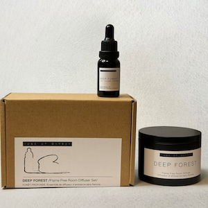 Może przedstawiać: Brązowe pudełko kartonowe z białą etykietą, na której widnieje napis "Deep Forest/Flame Free Room Diffuser Set/" oraz "Foret Profonde Ensemble de diffuseur d'ambiance sans flamme". Pudełko znajduje się obok czarnego słoika z białą etykietą, na której widnieje napis "Deep Forest Flame Free Room Diffuser" oraz "Diffuseur d'ambiance sans flamme". Na pudełku znajduje się mała czarna butelka z kroplomierzem i białą etykietą z napisem "Deep Forest".