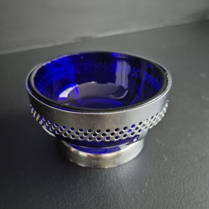 May include: A cobalt blue glass bowl with a silver metal rim and base. The rim features a decorative pattern of small, evenly spaced holes. The bowl's interior reflects light, highlighting its deep blue color. A decorative piece for the home.