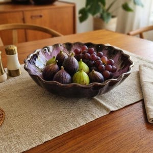 Peut inclure: Un bol en céramique à bords festonnés avec un vernis brillant violet et brun foncé, rempli d'une grappe de raisins violets frais, de plusieurs figues violettes foncées et de quelques figues vert clair, disposé comme centre de table.