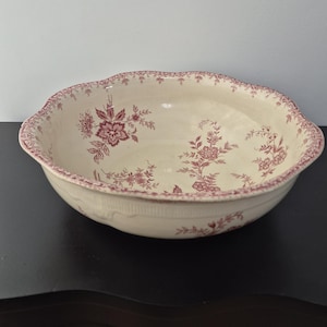 May include: A cream-colored, antique ceramic bowl with a scalloped edge and floral pattern in shades of burgundy. The bowl features a wide, shallow design, suitable for serving or display. The floral design is repeated around the rim and inside the bowl.