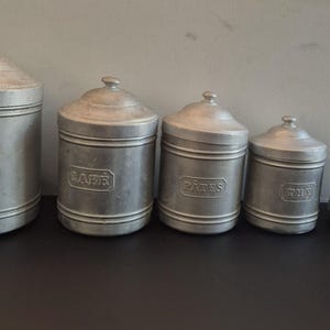 Op de afbeelding: Een set van vijf trapsgewijze cilindrische aluminium voorraadbussen met bijpassende deksels. Elke bus is voorzien van een horizontaal reliëflabel met de Franse woorden SUCRE, CAFE, PATES, THE en POIVRE op de voorzijde.