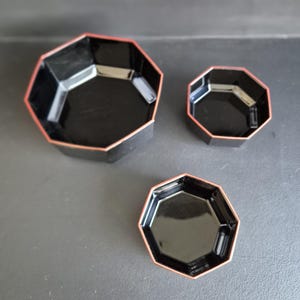 Peut inclure: Trois plats de service noirs octogonaux avec une bordure rouge. Les plats sont emboîtés, de tailles variées. La finition brillante reflète la lumière, soulignant les formes géométriques. Un design simple et élégant pour servir des amuse-gueules ou de petites portions.