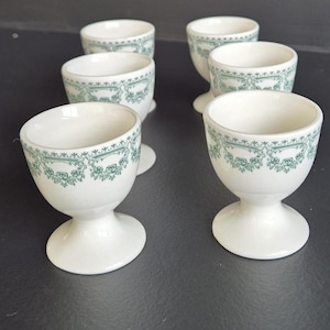 Peut inclure: Six coquetiers en céramique blanche avec un pied. Chaque tasse a un motif floral vert sur le bord. Les coquetiers sont disposés sur une surface sombre.