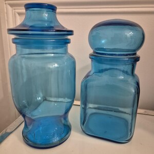 Op de afbeelding: Twee vintage blauwe glazen potten met deksels. Eén pot heeft een klassieke apothekersvorm, de andere is vierkant met een afgerond deksel. Beide potten zijn gemaakt van transparant blauw glas en worden waarschijnlijk gebruikt voor opslag.