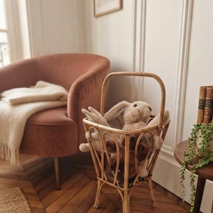 Puede incluir: Una cesta de mimbre trenzado de color marrón claro con un asa alta arqueada, apoyada sobre cuatro patas. La cesta contiene dos animales de peluche, un conejo beige claro y un oso marrón oscuro, colocados dentro de la estructura abierta.
