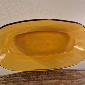 Puede incluir: Un plato de vidrio de forma ovalada y color ámbar, con un borde marrón oscuro. El plato tiene una superficie texturizada y probablemente se utiliza para servir alimentos o como pieza decorativa. El vidrio parece transparente.