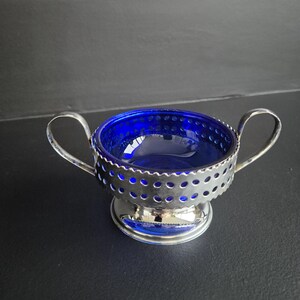 May include: A vintage sugar bowl with a vibrant cobalt blue glass interior and a silver-toned metal filigree exterior. The bowl has two curved handles and sits on a small, round base. The design is elegant and classic.