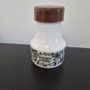 Peut inclure: Un pot à moutarde en céramique blanche avec un couvercle marron. Le pot présente un motif floral noir et les mots "Moutarde Mosterd". Le pot est de forme arrondie et est probablement vintage.