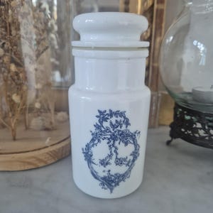May include: A white milk glass apothecary jar with a cylindrical body and a matching lid. The front of the jar features a detailed blue transferware print depicting a floral wreath design with intertwined vines, leaves, and small blossoms.