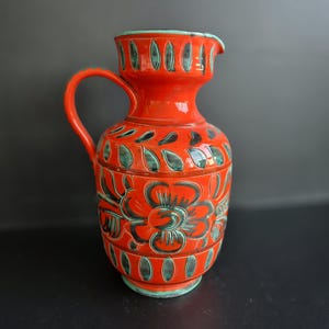 May include: A vibrant, red ceramic pitcher with a handle and spout. The pitcher is adorned with a floral design and leaf-shaped accents in teal and black. The rim and base are also teal. A decorative piece for the home.
