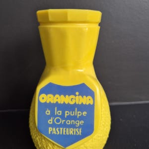 Può includere: Una bottiglia vintage di Orangina gialla con una superficie strutturata. La bottiglia ha un'etichetta blu con testo bianco che recita "ORANGINA à la pulpe d'Orange PASTEURISE". La bottiglia ha un tappo giallo.