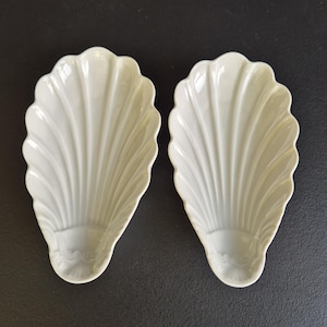 Può includere: Due piatti in ceramica color bianco sporco a forma di conchiglie allungate. Ogni piatto presenta una serie di nervature in rilievo che si irradiano da una base arrotondata verso un bordo smerlato, creando un aspetto testurizzato a ventaglio.