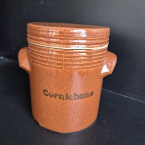 Peut inclure: Un pot en céramique marron avec un couvercle, portant le mot "Cornichons" en écriture cursive. Le pot a une surface texturée avec des rainures horizontales et de petites poignées de chaque côté. Le pot est conçu pour conserver les cornichons.