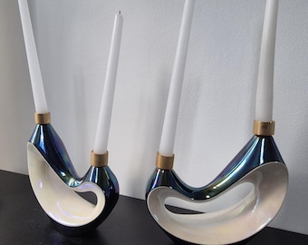 May include: Two decorative candle holders with white candles. The holders have a dark blue, iridescent finish with a white, curved design. The candle holders have a gold-colored base and are placed on a black surface.