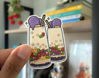 May include: A die-cut vinyl sticker featuring two tall glasses of bubble tea. Each glass is filled with a beige liquid, black tapioca pearls, and colorful red and green jelly cubes. A scoop of purple ice cream and a striped straw top each drink.