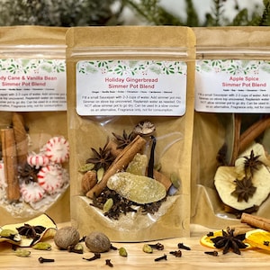 May include: Three kraft paper bags filled with simmer pot blends. The bags are labeled "Candy Cane & Vanilla Bean", "Holiday Gingerbread", and "Apple Spice". Each bag contains a mix of spices, herbs, and dried fruit. The bags are displayed on a wooden surface.