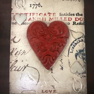 May include: A rectangular, aged-looking decorative piece with a large, textured red heart in the center. The background features faded text, the year "1776", and the word "LOVE". There are also white heart-shaped embellishments.