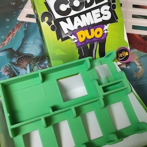 May include: A green game board with a cutout for a game card. The game board is for the game Code Names Duo, a two-player version of the popular word association game.