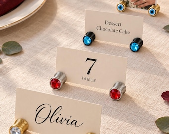 May include: Four sets of cylindrical metal place card holders with decorative ends. The holders feature clear, red, blue, and evil eye designs, supporting white cards printed with 'Olivia', '7 TABLE', 'Dessert Chocolate Cake', and a photo.
