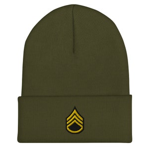 May include: Olive green beanie with a Sergeant rank insignia embroidered in gold and black. The beanie is a knit cap with a folded brim, suitable for cold weather. The Sergeant rank insignia is a chevron design.