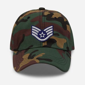 May include: Camouflage baseball cap with a military insignia patch. The cap features a classic six-panel design with a curved brim. The patch is a blue and white emblem with a star in the center. The cap is made of a durable fabric.