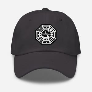 Puede incluir: Gorra de béisbol gris carbón con el logotipo blanco y negro de la Iniciativa Dharma en la parte delantera. El logotipo es un octágono estilizado con la palabra "DHARMA" encima de un símbolo curvo. La gorra tiene una visera curva y una correa ajustable.