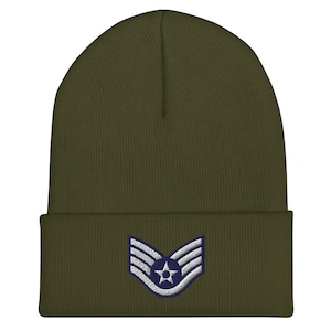 May include: Olive green beanie with a folded brim. The front features an embroidered insignia with a star and stripes in white and navy blue. The beanie is made of a soft, knitted material.