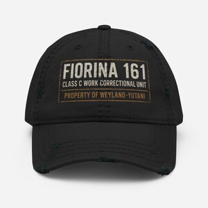 Op de afbeelding: Zwarte, versleten baseballcap met de tekst "FIORINA 161 CLASS C WORK CORRECTIONAL UNIT PROPERTY OF WEYLAND-YUTANI" in een rechthoekig ontwerp. De cap heeft een gebogen klep en een versleten uiterlijk.