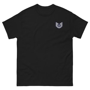 Peut inclure: T-shirt noir avec un petit écusson brodé sur la poitrine gauche. L'écusson présente une étoile au-dessus de trois bandes, dans un coloris bleu clair et blanc. Le t-shirt a un col rond et des manches courtes.