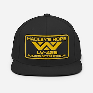Puede incluir: Gorra de béisbol negra con visera plana y parche rectangular amarillo. El parche presenta el texto "HADLEY'S HOPE" sobre un logotipo "W" estilizado, "LV-426" y "BUILDING BETTER WORLDS" en letras amarillas.