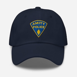 Pode incluir: Um boné de beisebol azul-marinho com um emblema bordado na frente. O emblema é um triângulo azul com borda amarela, contendo o texto amarelo AMITY POLICE e uma pequena forma de diamante amarela na ponta inferior do triângulo.