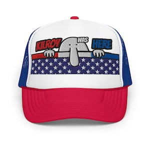 Könnte beinhalten: Eine weiße und rote Trucker-Kappe mit blauem Netzrücken. Die Kappe zeigt ein gesticktes Bild eines Comic-Charakters, der über einer rot-weiß-blauen amerikanischen Flagge mit dem Text "Kilroy Was Here" lugt.