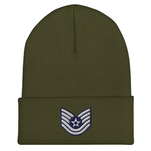 Peut inclure: Bonnet vert olive avec un bord replié. L'avant présente un écusson avec une étoile et des rayures blanches sur fond bleu marine. Le bonnet est en maille douce.